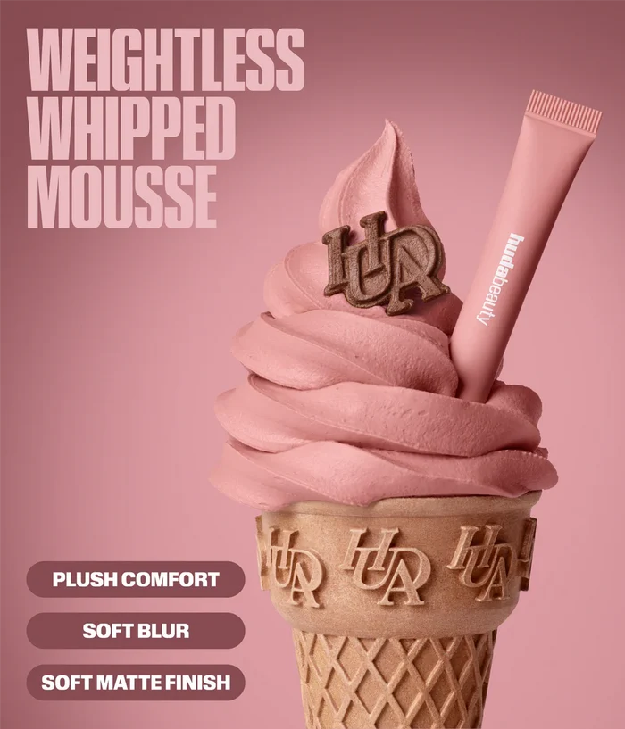 A pink lip product tube labeled 'hudbeauty' shown on top of a pink whipped mousse swirled in a waffle cone. Large text reads 'WEIGHTLESS WHIPPED MOUSSE' with marketing callouts 'PLUSH COMFORT', 'SOFT BLUR', and 'SOFT MATTE FINISH'.